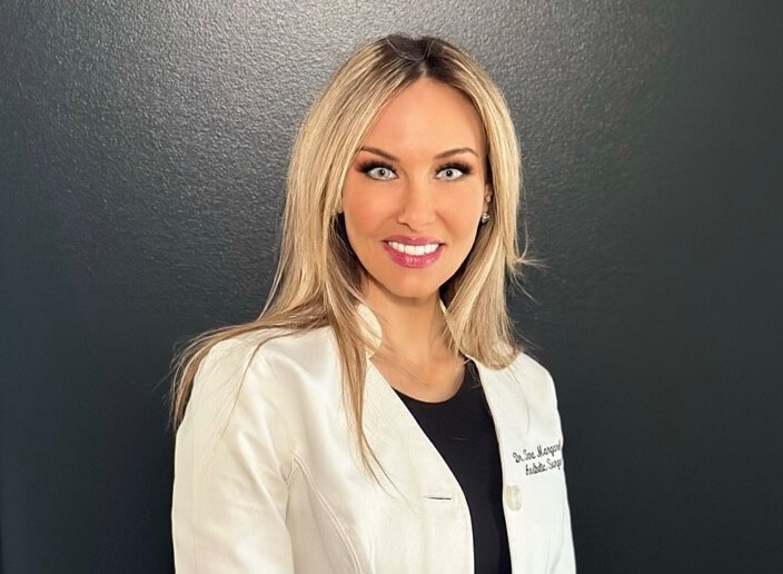 Portrait of a smiling woman with blonde hair wearing a white lab coat, standing against a dark background.